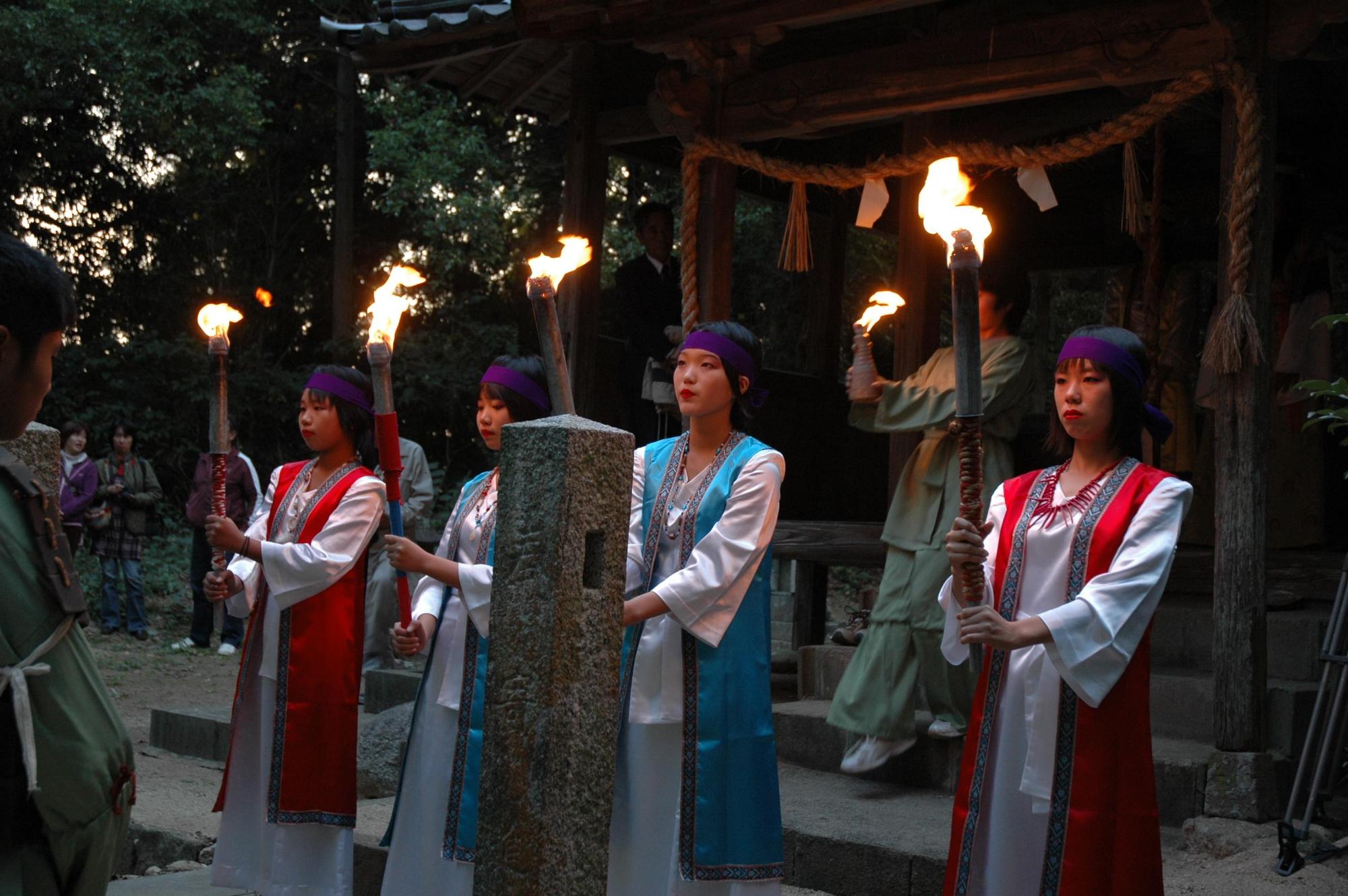 白地に赤色と青色の袖のない羽織を着た女性4名が火のついたたいまつを持って神社の社の前に立っている写真