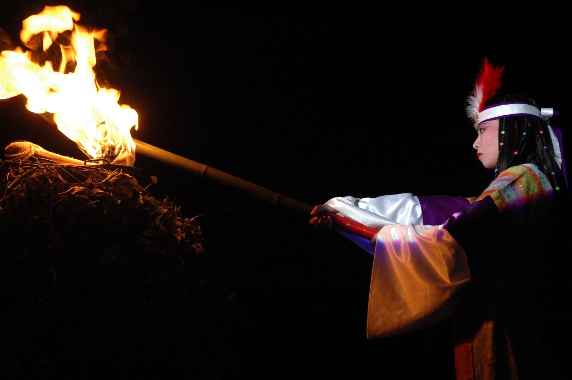 卑弥呼の衣装を身に着けた女性が炎の付いたたいまつを持って火を付けようとしている写真