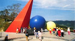 展望台にシンボルマークとして赤い三角すい、青い球、少し小さい黄色い球が設されており、子ども達がシンボルマークの周りに集まっている写真