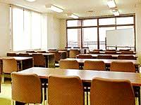 ホワイトボードが前に置かれ、長い机に椅子が3個ずつ並んでいる学習室の写真